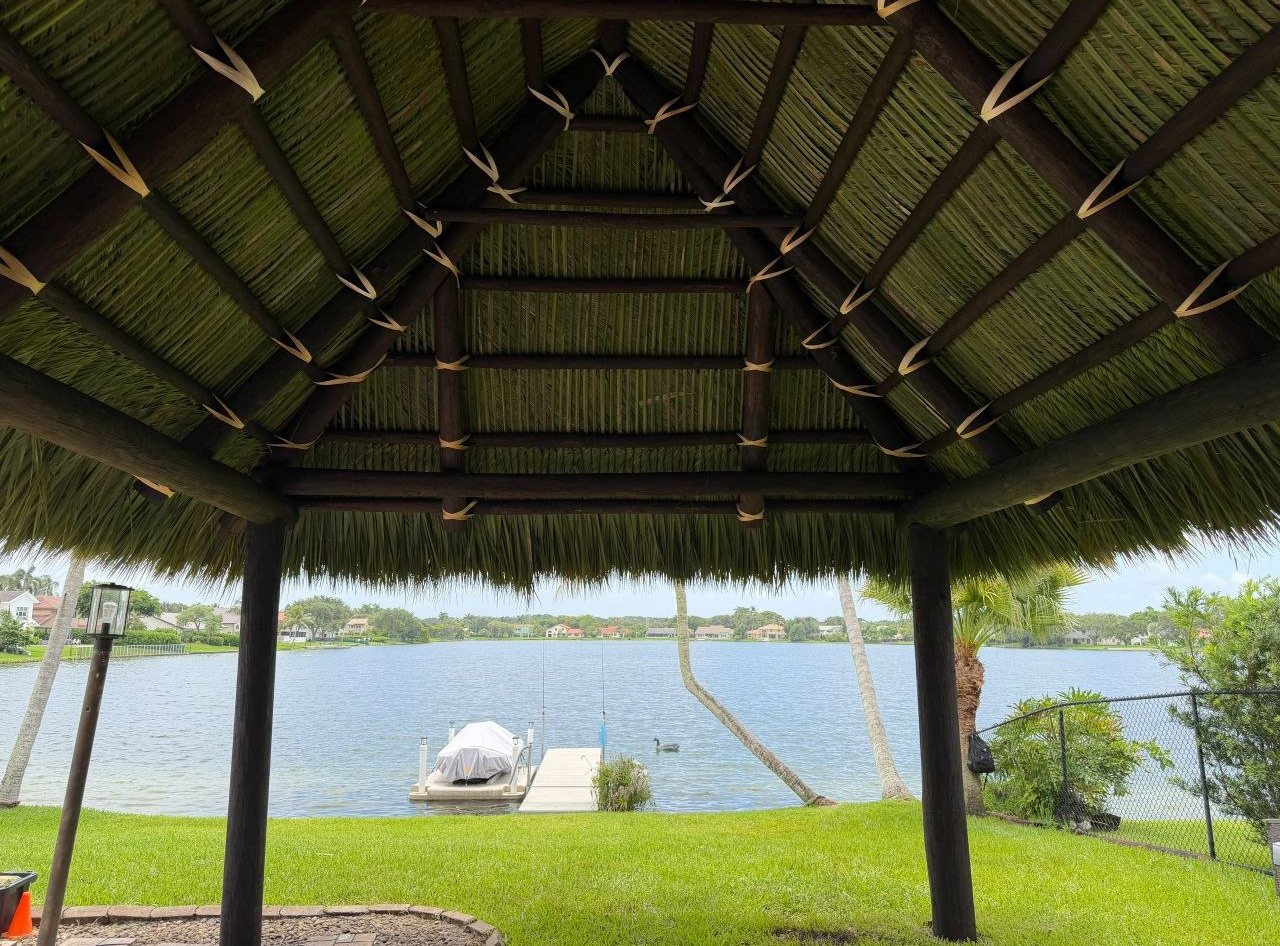 View from under a thatched tiki hut out over a lake and dock. Mai Tiki Huts builds from the Panhandle to the Keys.